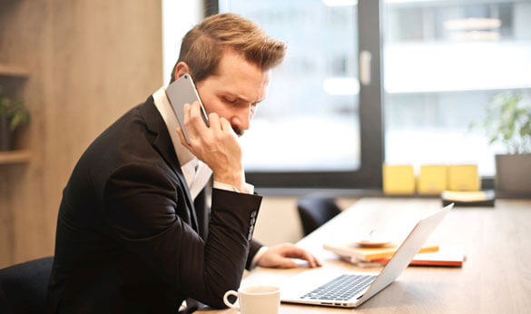 A man is sitting in the office with his laptop open, holding his mobile to his ear while listening to